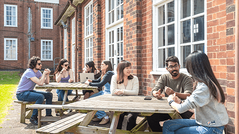 Two wooden tables outside Harry French student accommodation full of students laughing and talking together in the sunny weather