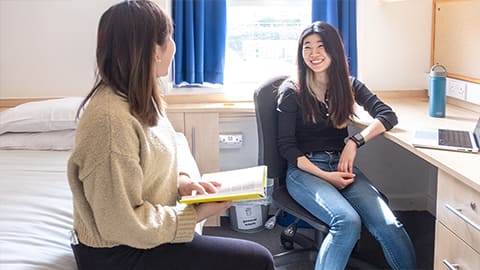 Two students chatting and laughing in a spacious bedroom in John Phillips, one student is sat reading on the bed and another is working at her desk