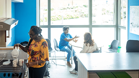 Students in the John Phillips kitchen, one is using the kettle in the kitchen and another two students are sat round the dining table chatting together