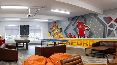 Recently renovated common room in Rutherford Loughborough student halls with bright orange bean bags, brown sofas, two drinks bars, table games and a painted mural on the wall