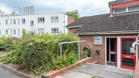 A daytime exterior view of the Telford Reception building, featuring a single‑storey brick entrance with a red door and a sign reading “Telford Reception.” The path leading to the door is bordered by metal handrails and surrounded by greenery, with a larger white accommodation building visible in the background under a cloudy sky.