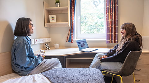 Students chatting together in a student bedroom in the Holt student accommodation. One student is working sat at her desk and the other student is chatting to her from the bed.