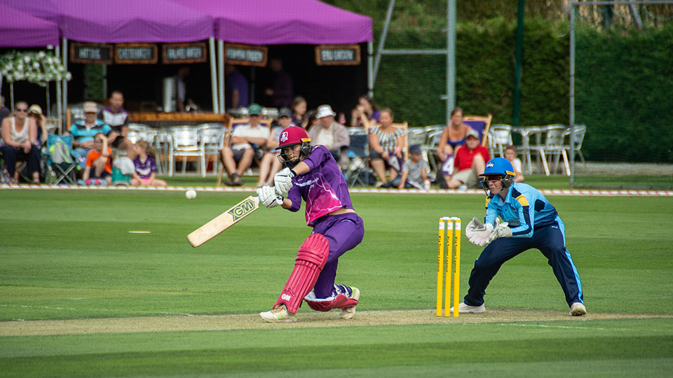 Two female crickets - one holding a bat hitting and ball and one fielding