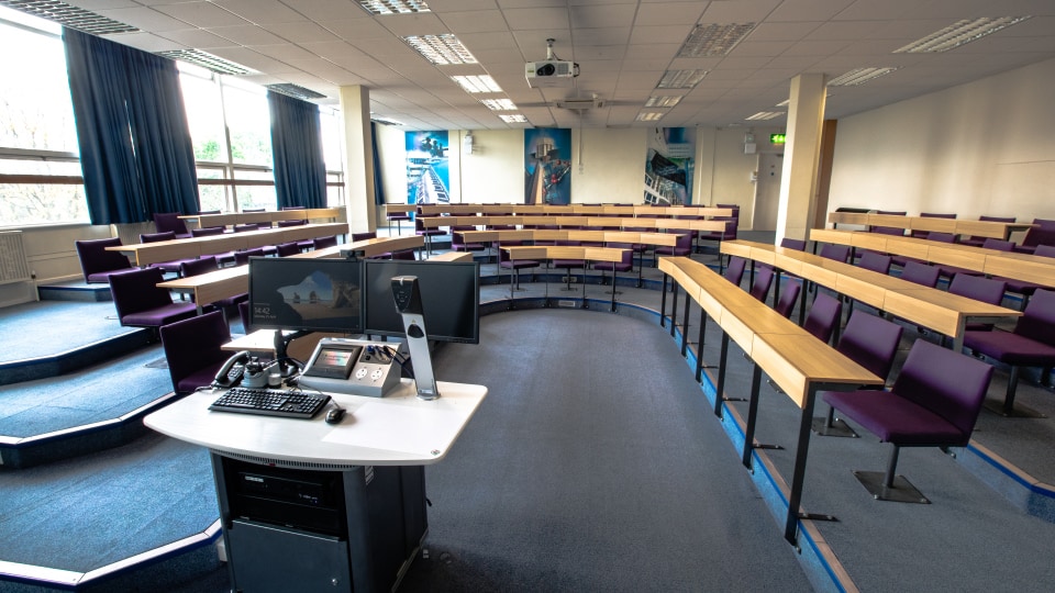 Brockington B111 - Lecture theatre with tiered curved seating, fixed desks, large projection screen at front, and ceiling lighting.