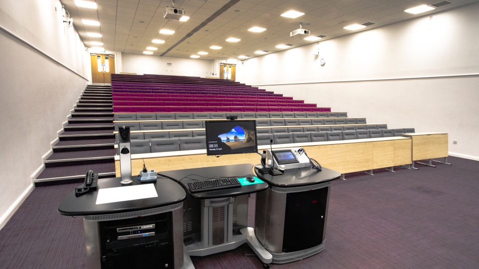 James France CC011 - Lecture hall with tiered seating, purple and gray chairs, and a central podium equipped with computer and microphone.
