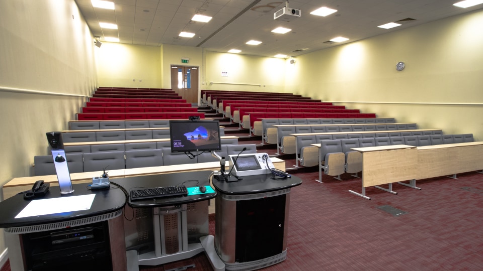 James France CC013 - Lecture hall with tiered red and gray seating, projector, and lectern equipped with computer and microphone.