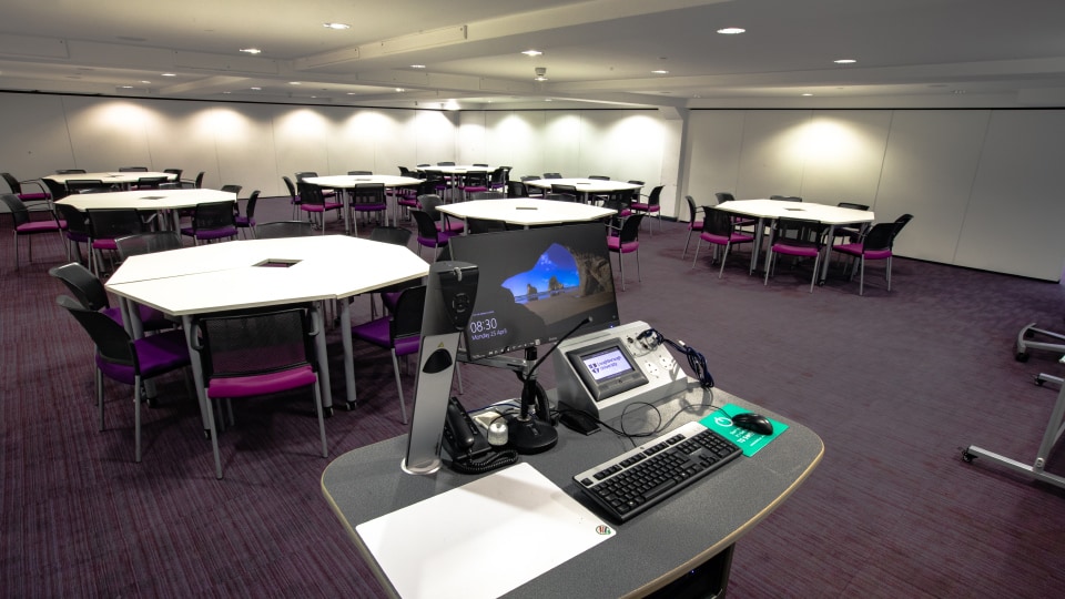 James France CC029a - Modern teaching room with hexagonal tables, purple chairs, and a presentation podium equipped with a computer and microphone.