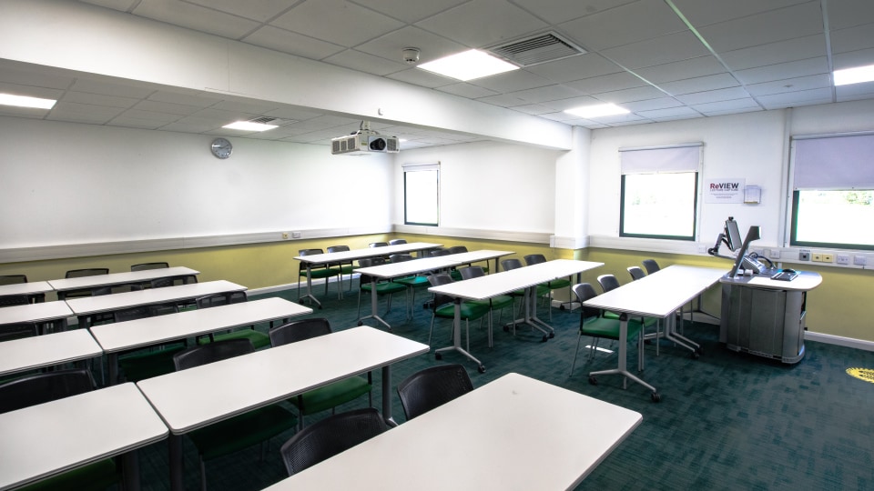 James France CC110 - Spacious teaching room with rows of white tables, green chairs, projector, and windows.
