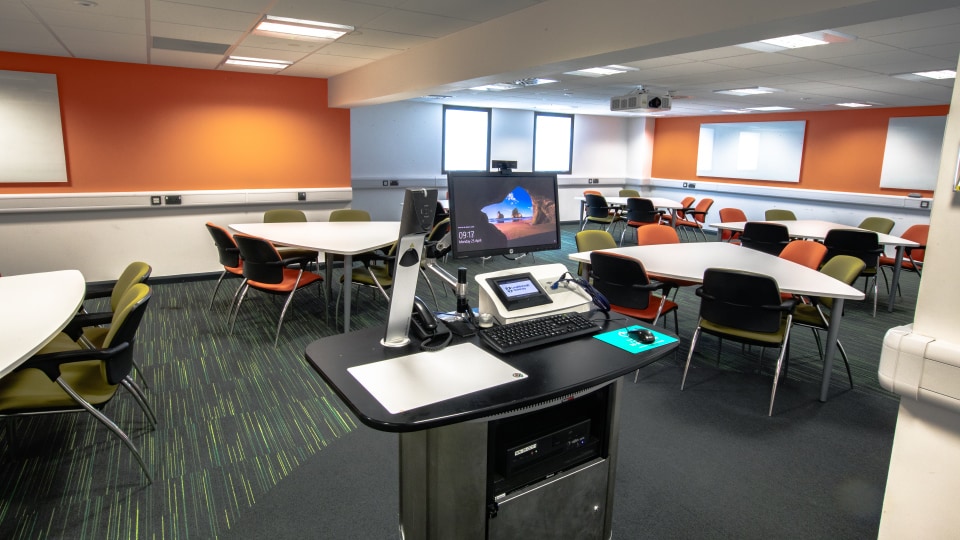 James France CC111 - Modern classroom with white tables, green chairs, projector, and computer station. Brightly lit with windows and blinds.