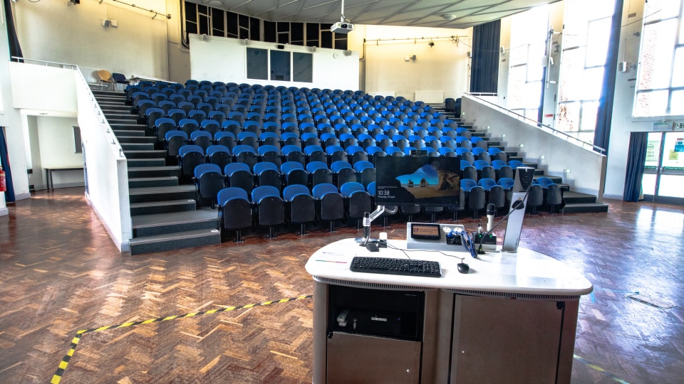 Cope auditorium - Large lecture theatre with tiered seating in a row, central moveable projection screen, large open space and ceiling lighting.