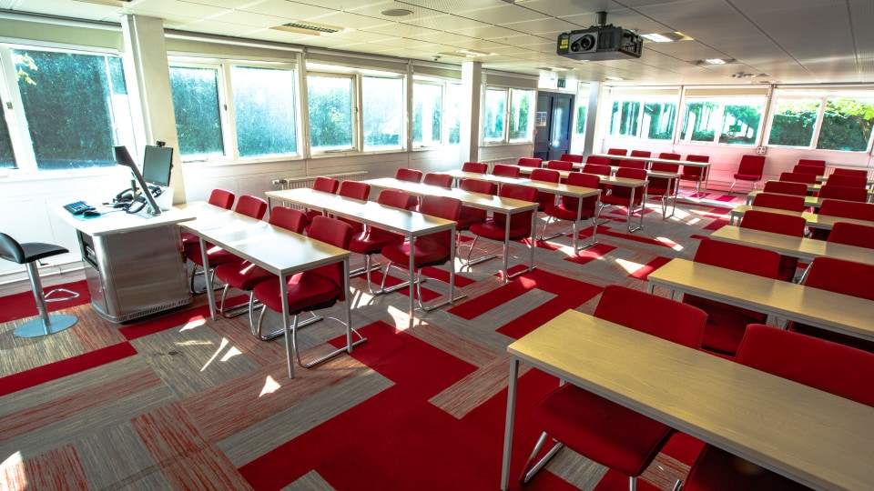 James France D002 - Bright classroom with red chairs, wooden desks, large windows, and a ceiling-mounted projector.