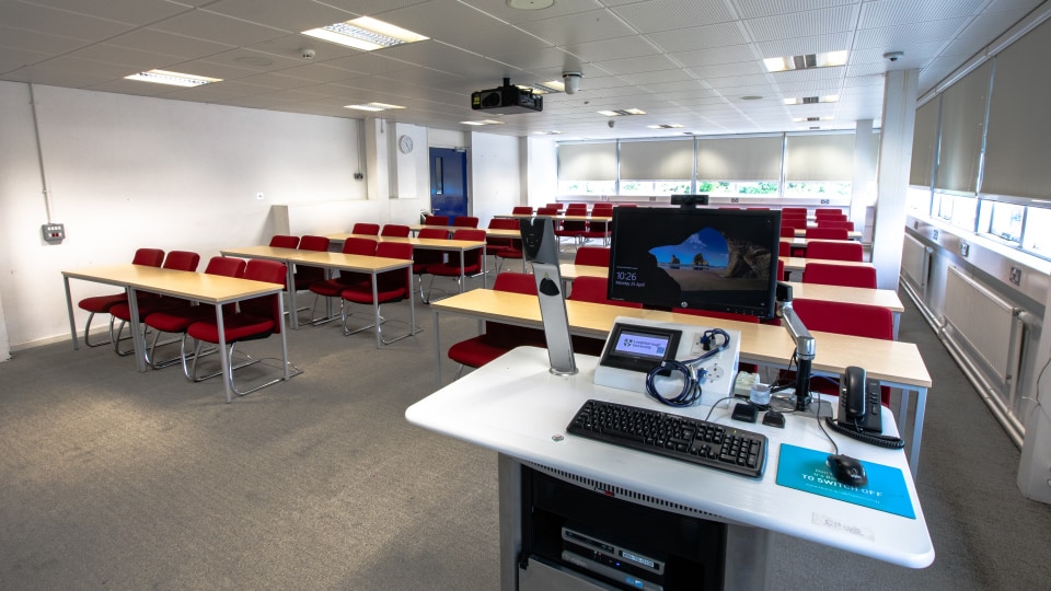 James France D102 - Modern teaching room with red chairs, wooden desks, and a multimedia podium featuring a computer and control panel.