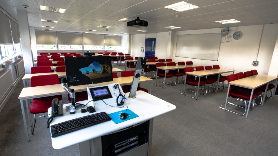 James France D109 - Teaching room with red chairs, wooden desks, projector, and instructor's podium with computer and phone.