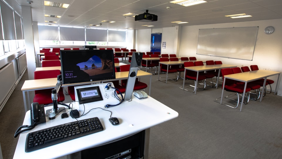 James France D201 - Modern teaching room with red chairs, tables, and a lectern equipped with computer and microphone. Large windows and whiteboard.