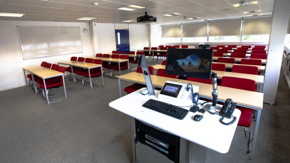 James France D202 - Teaching room with rows of red chairs, wooden desks, and a lectern featuring a computer and microphone.