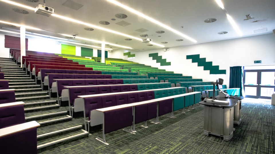 EHB 104 - Lecture hall with tiered seating in purple and green, projector, and podium. Bright lighting and modern design.