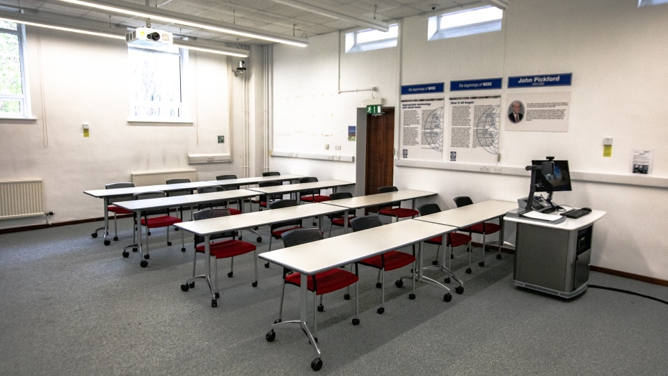 John Pickford HH022 - Teaching room with rows of tables, red chairs, projector, and computer station. Posters on walls, windows above.