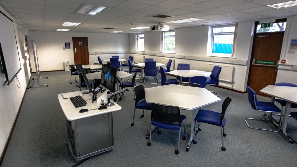 John Pickford HH023 - Teaching room with hexagonal tables, blue chairs, computer workstation, projector, and whiteboard. Bright, spacious environment.