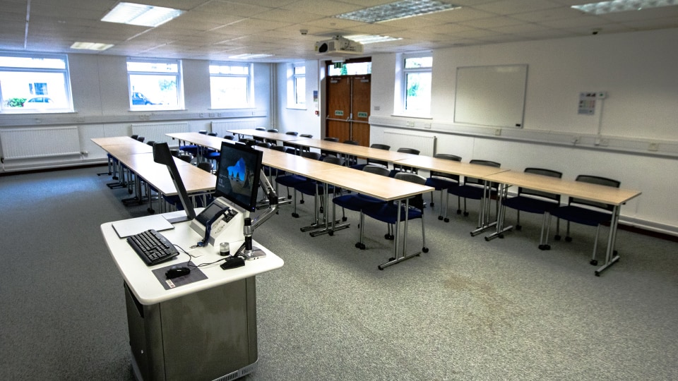 John Pickford HH023a - Teaching room with rows of tables, chairs, a computer podium, whiteboard, and windows.