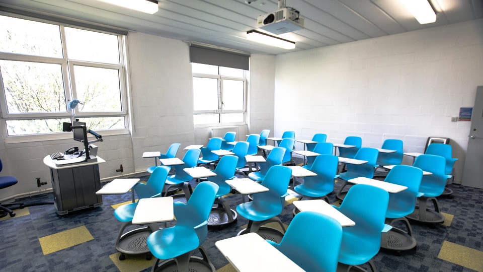 Matthew Arnold MAR103 - Teaching room with blue swivel chairs, attached desks, large windows, and a podium with computer equipment.