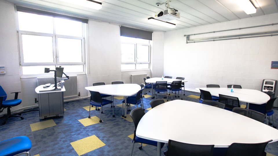 Matthew Arnold MAR105 - Teaching room with triangular tables, chairs, projector, and computer station. Blue and yellow checkered carpet.