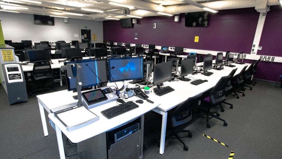 S Building S006 - Computer lab with rows of desks, monitors, keyboards, and chairs. Purple walls and overhead lighting.