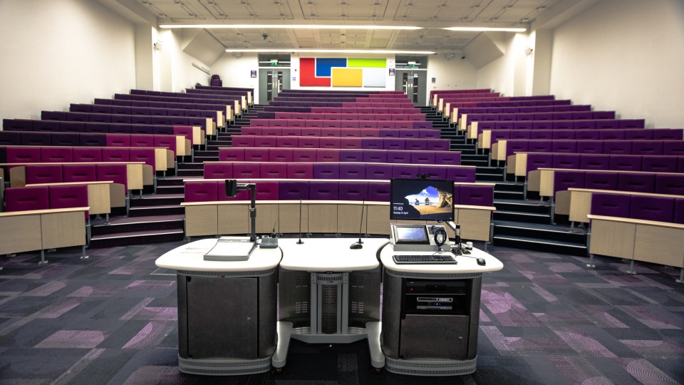 Wolfson T003 - Lecture theatre with tiered purple seating, central podium with computer, microphone, and projector. Colourful wall panels at back.