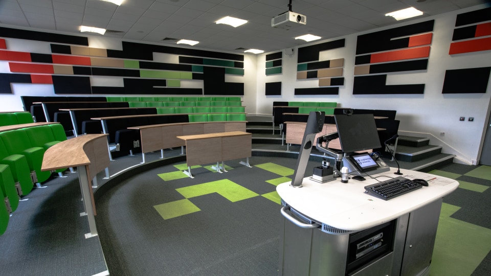 Brockington extension U005 - Lecture theatre with tiered seating, fixed desks, large projection screen at front, and ceiling lighting.