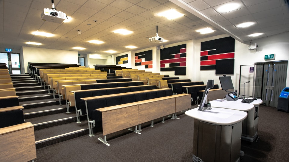 Brockington extension U020 - Large lecture theatre with tiered seating, fixed desks, dual projection screens at front, and ceiling lighting.