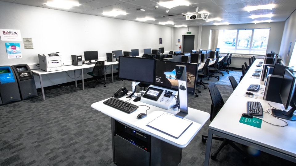 Wavy Top WAV056 - Computer lab with rows of desks, monitors, keyboards, and a central teaching station. Bright lighting, large window, and wall posters.