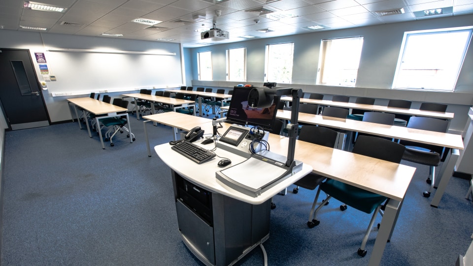 Wavy Top WAV120 - Teaching room with rows of desks and chairs, equipped with a lectern featuring a computer, keyboard, and projector.