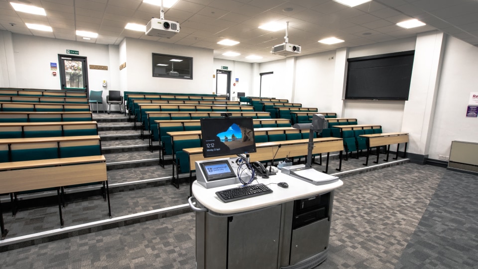 Wavy Top WAVB01 - Lecture theatre with tiered seating, green chairs, wooden desks, and a central podium featuring a computer and projector screen.