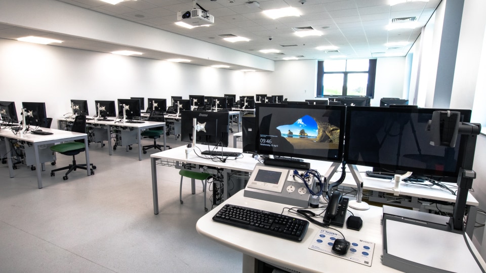 West Park Labs WPL105 - Spacious computer lab with rows of desks, each equipped with monitors and chairs. A control station with dual screens in foreground.