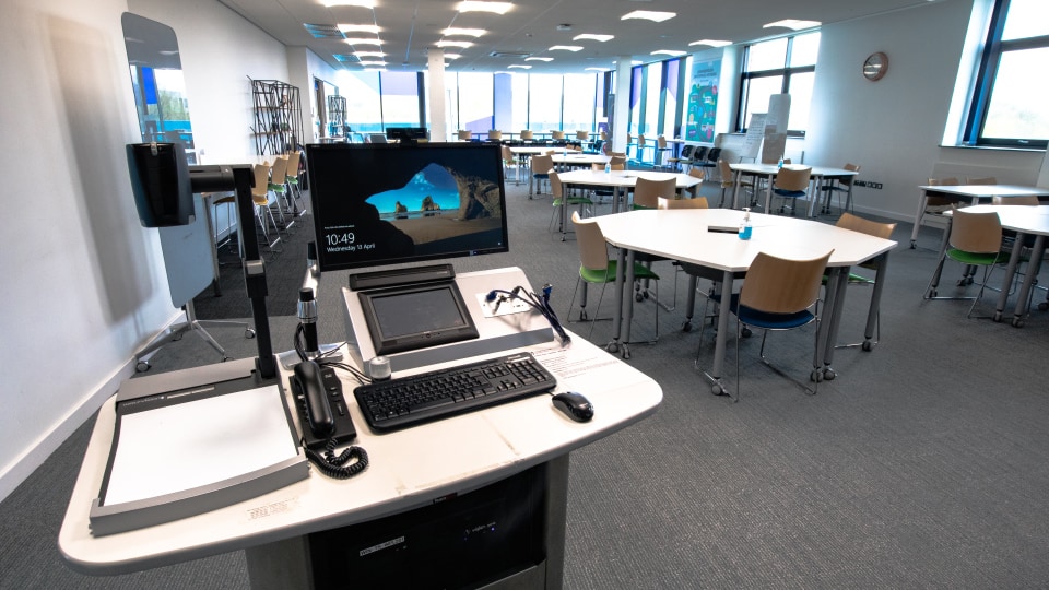 West Park Labs WPL201 - Teaching room with modern lectern, computer, and projector. Multiple tables and chairs arranged for collaborative learning. Bright windows.
