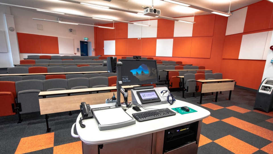 West Park Teaching Hub WPT001 - Lecture theatre with tiered seating, orange and gray color scheme, and a central podium featuring computer equipment and controls.