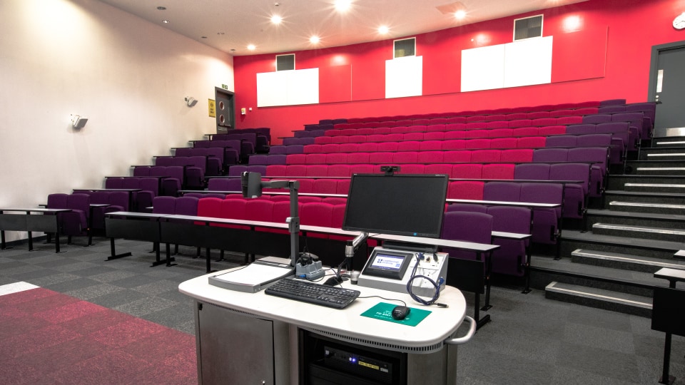 West Park Teaching Hub WPT003 - Lecture theatre with tiered seating in red and purple, a lecturer's desk with computer and equipment, and red accent wall.