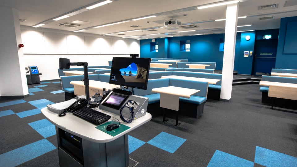 West Park Teaching Hub WPT005 - Modern lecture theatre with tiered blue seating, individual desks, and a central teaching podium equipped with computer and audio-visual controls.