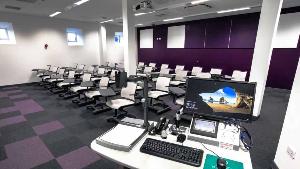 West Park Teaching Hub WPT006 - Modern teaching room with rows of swivel chairs and desks, purple and gray carpet, and a technology-equipped instructor's podium.