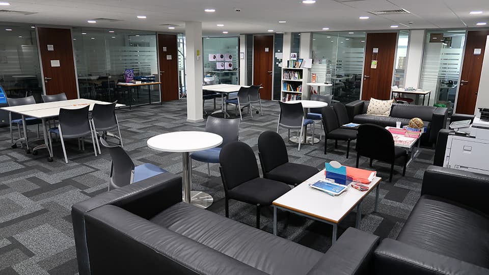 Teaching Room HH024 - Spacious teaching room with modern furniture, including sofas, round tables, chairs, and bookshelves. Glass walls separate additional rooms.