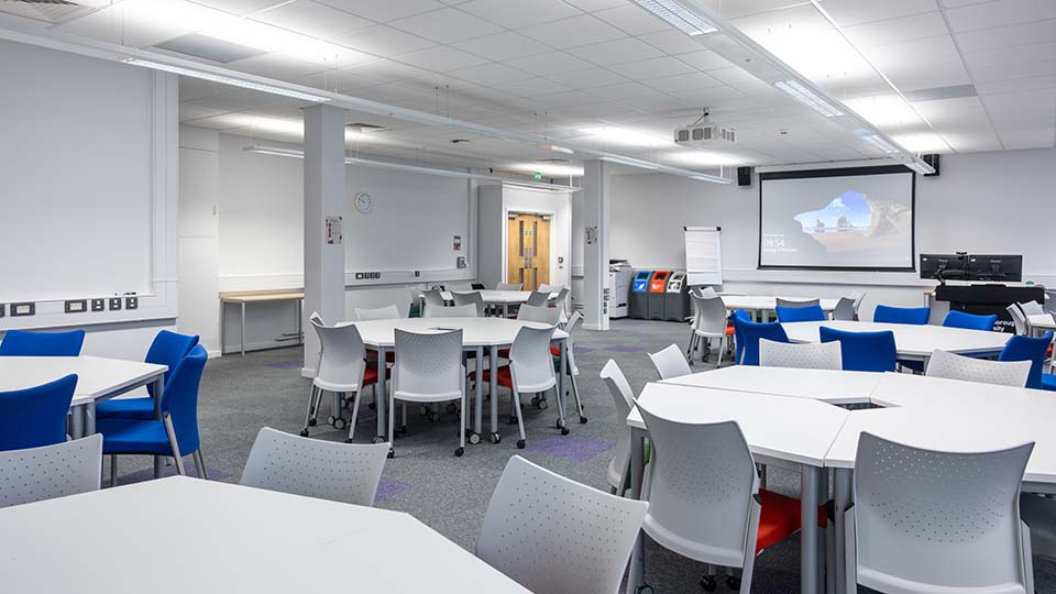 B007-teaching-room - Teaching room with flat layout, grouped tables, projector screen at front, white walls, and ceiling lights.