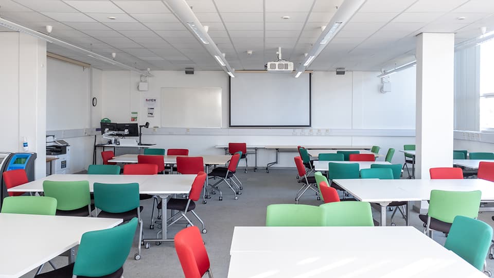 b114-teaching-room - Teaching room with flat layout, grouped desks, projector screen and whiteboard at front, bright lighting.