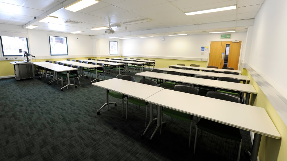 James France CC109 - Spacious teaching room with rows of white tables, green chairs, projector, and windows.