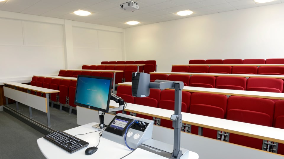 Edward Barnsley EB63107 - Small lecture theatre with tiered seating, fixed desks, projection screen at front, and ceiling lighting.