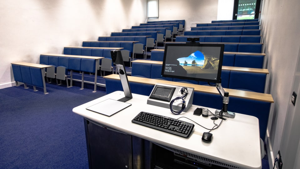 David Davies DAV028 - Lecture theatre with tiered blue seating, a teaching podium featuring a computer, keyboard, mouse, and digital display screen.