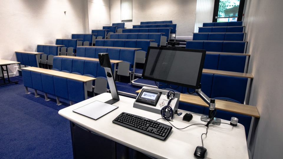 David Davies DAV029 - Lecture theatre with tiered blue seating, a teaching podium featuring a computer, keyboard, mouse, and digital display screen.