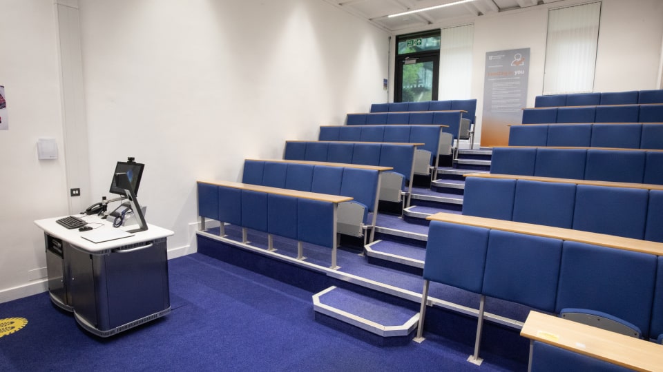 David Davies DAV030 - Lecture theatre with tiered blue seating, a lectern with computer and microphone, and a poster near the exit door.