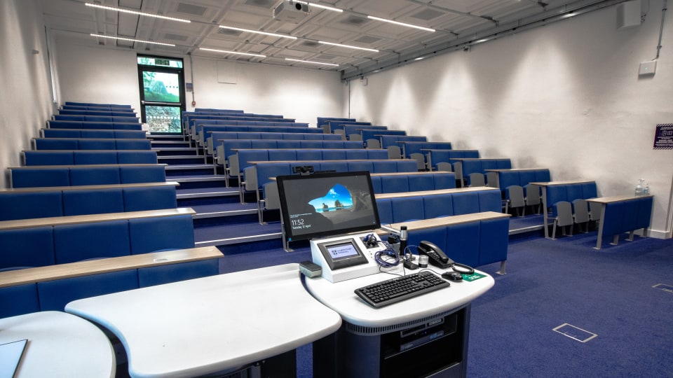 David Davies DAV031 - Lecture theatre with tiered blue seating, white walls, and a teaching podium equipped with a computer and control panel.