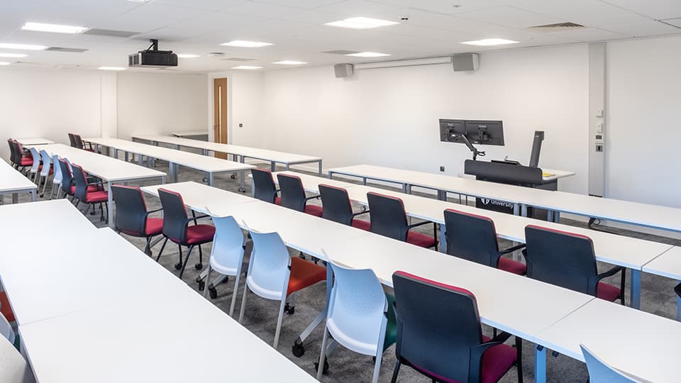 David Davies Dav1064 - Modern teaching room with long white tables, colourful chairs, dual monitors on a podium, and ceiling projector.