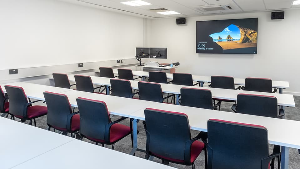 David Davies DAV1065 - Teaching room with rows of desks and chairs facing a large screen displaying a beach scene. Dual monitors on a podium.