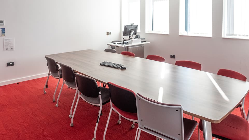 David Davies DAV1095 - Teaching room with a long table, eleven chairs, red carpet, and a computer station near windows. Bright, natural light.